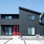 片流れ屋根と黒い外壁、赤い玄関ドアを組み合わせた2階建て住宅